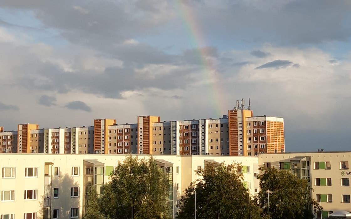 Ein schwacher Regenbogen erscheint am bewölkten Himmel über Wohnhäusern und Bäumen in Rostock.