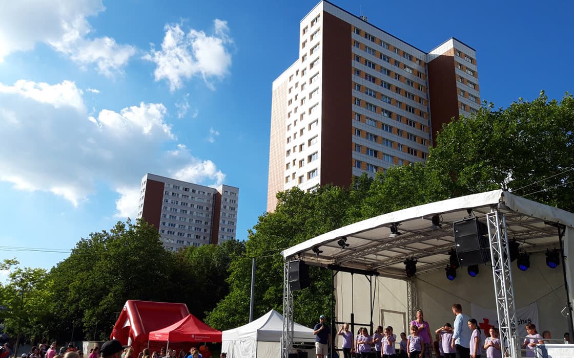 Menschen versammeln sich bei einer Freiluftveranstaltung in Rostock, mit einer Bühne, Zelten und hohen Wohnhäusern im Hintergrund.