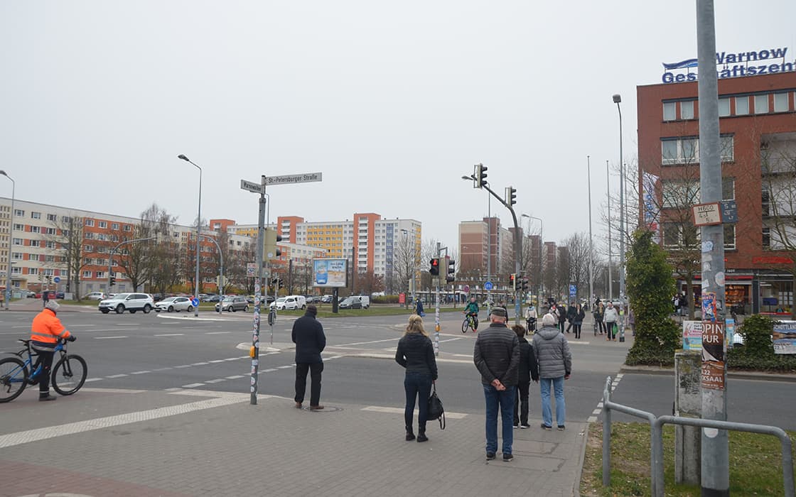 An einem bewölkten Tag warten Menschen an einem Zebrastreifen in Rostock, einem städtischen Gebiet mit Wohnungen, Autos und Fahrrädern.