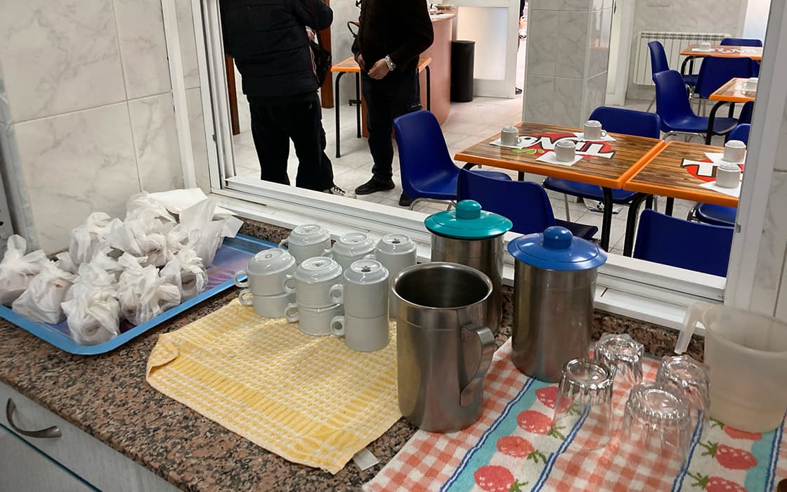 Eine Theke mit Tassen, Thermoskannen, Gläsern und eingepackten Lebensmitteln in Misión Urbana in Valencia; blaue Stühle im Hintergrund.
