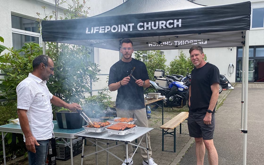Drei Männer grillen Fleisch unter einem Vordach der Lifepoint-Kirche in Stuttgart, Rauch steigt vom Grill auf.