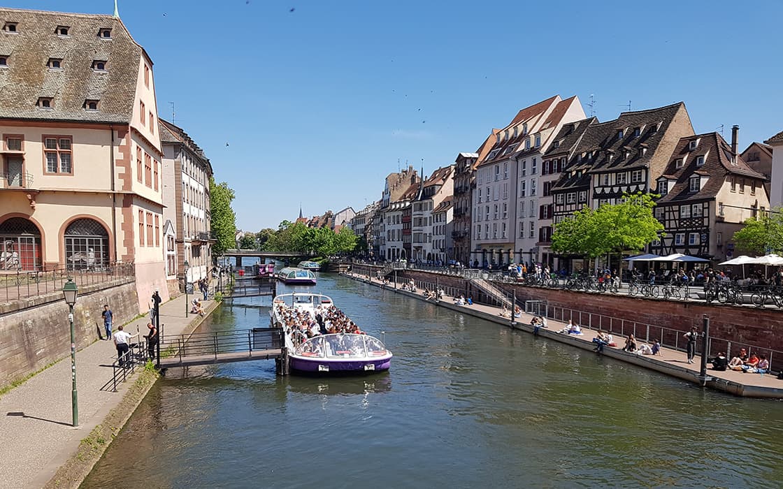 Ein Ausflugsboot auf einem Kanal fährt unter blauem Himmel in einer europäischen Stadt an Gebäuden aus der Zeit der Gemeindegründungen vorbei.