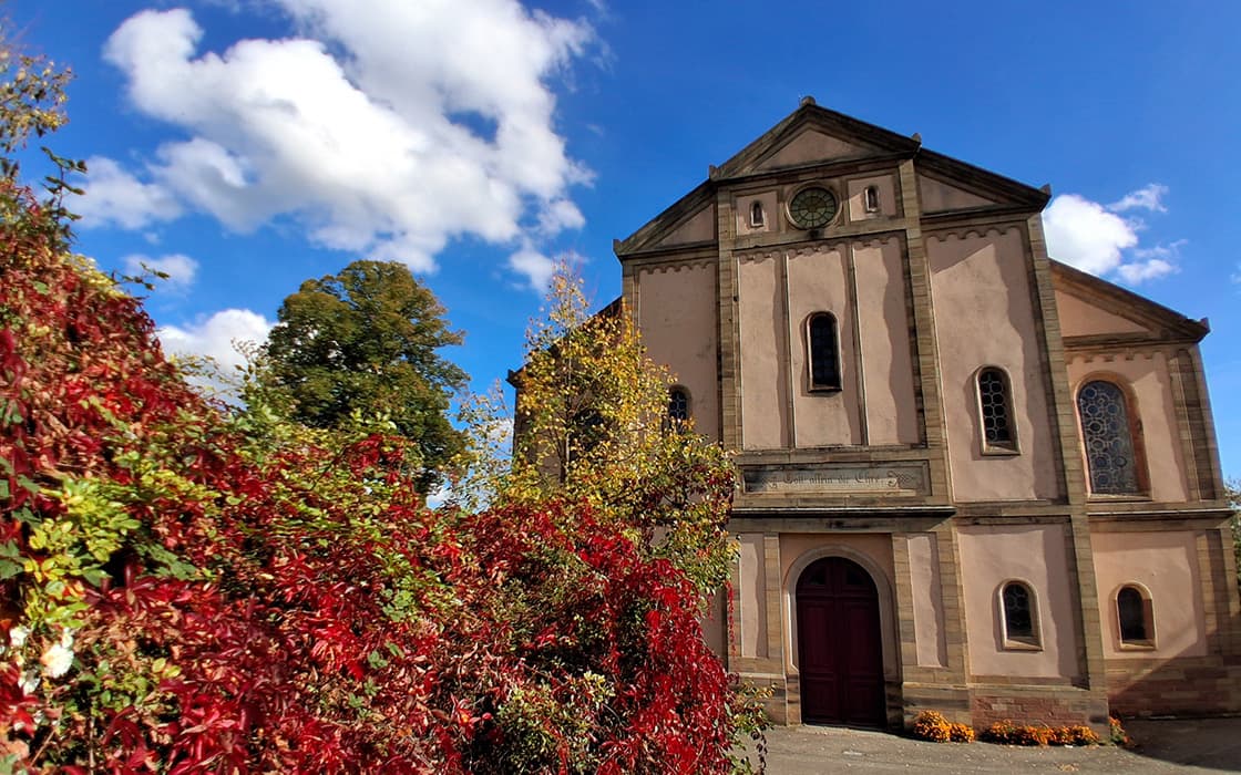 Historische Kirche in Westfrankreich mit Rundbogenfenstern, roter Tür und Herbstlaub unter strahlend blauem Himmel.