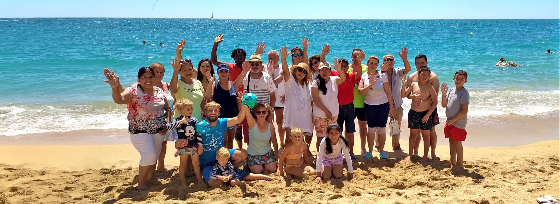 Eine Gruppe von Menschen, darunter Kinder und Erwachsene, lächeln und winken am Sandstrand der Costa Azahar in Spanien.