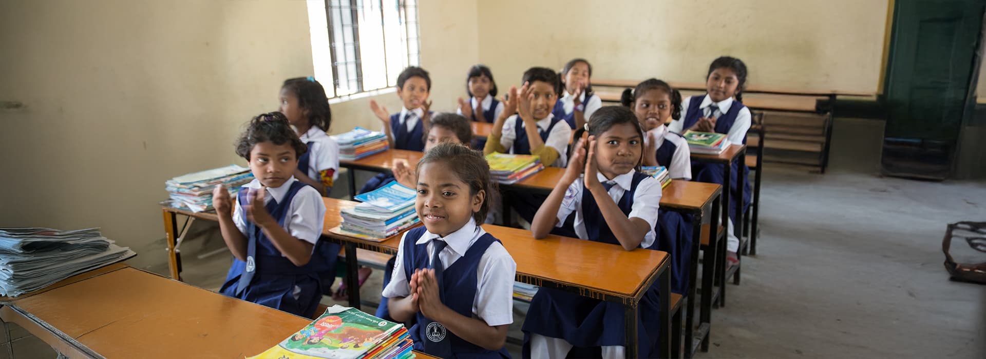 Junge Schüler klatschen in einem hellen Klassenzimmer des Kinderdorfs Dinajpur in Bangladesch, die Bücher liegen fein säuberlich gestapelt auf ihren Tischen.