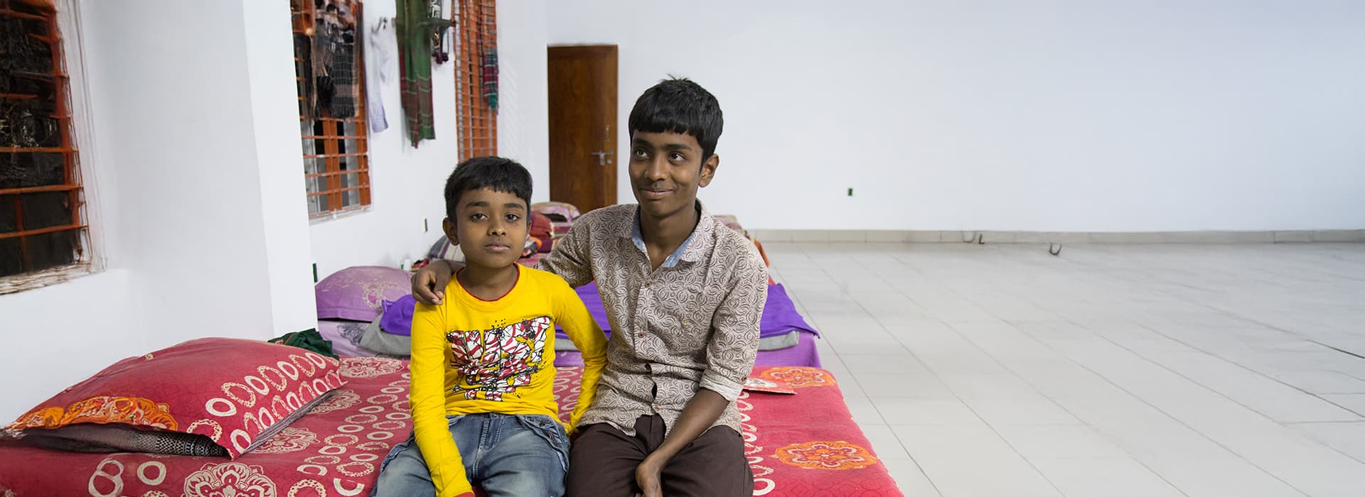 Zwei Jungen sitzen zusammen auf einem Bett in einem hellen, farbenfrohen Zimmer im Kinderdorf Khulna in Bangladesch.