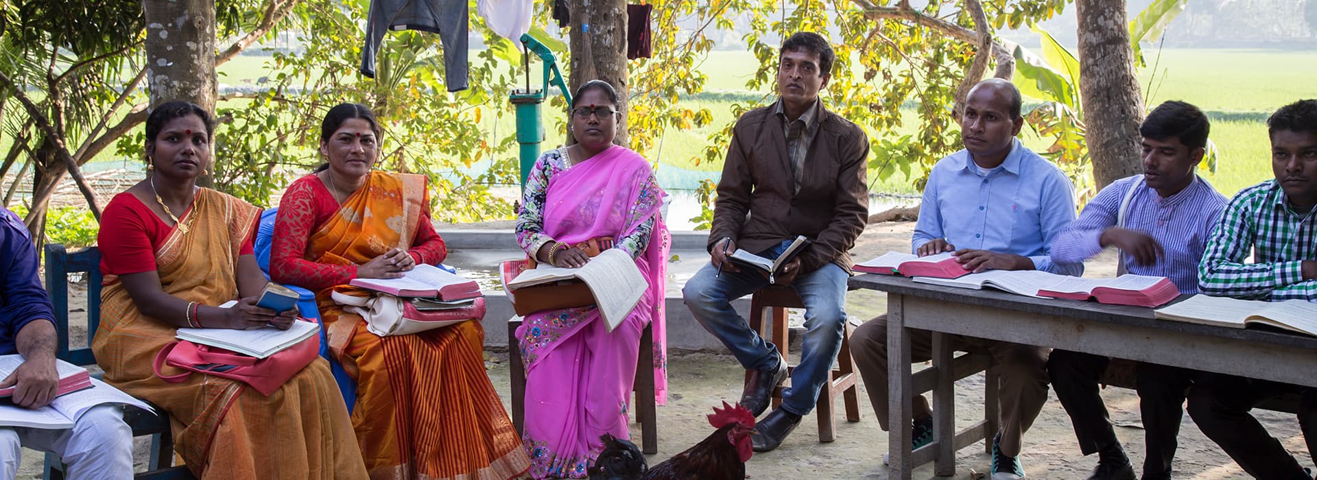 Eine Gruppe versammelt sich im Freien im Kreis zur theologischen Ausbildung, Bücher in der Hand, Bäume und Felder im Hintergrund.