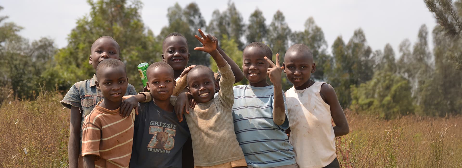Eine Gruppe von sieben lächelnden Kindern steht in Burundi im Freien vor hohem Gras und Bäumen zusammen.