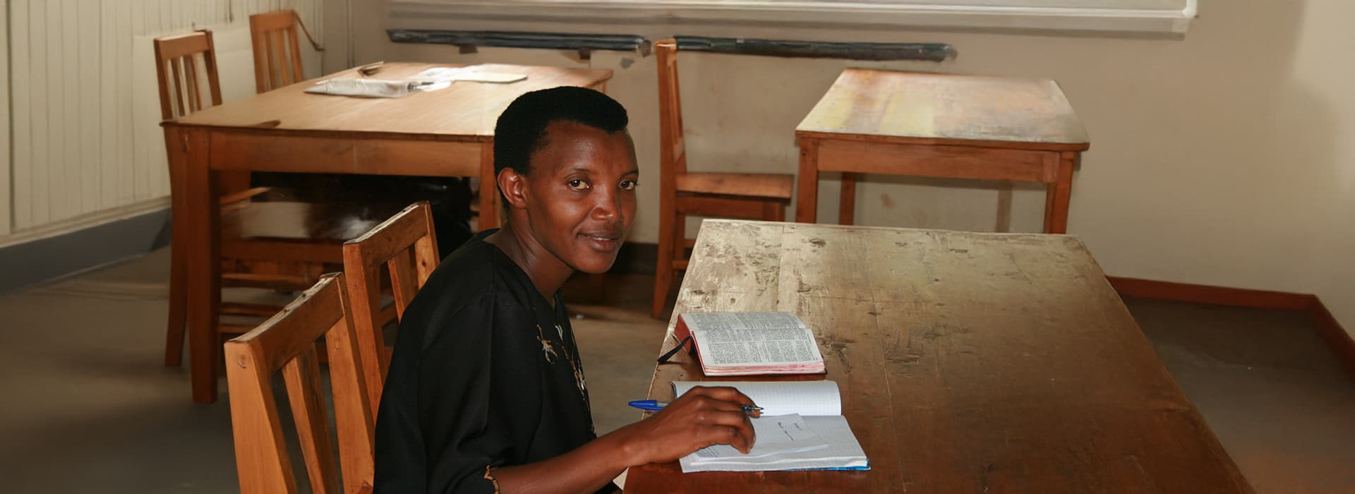 Eine Frau sitzt allein an einem Holztisch in der Bibelschule Muramvya, Burundi, und liest und schreibt in einem sonnenbeschienenen, ruhigen Raum.