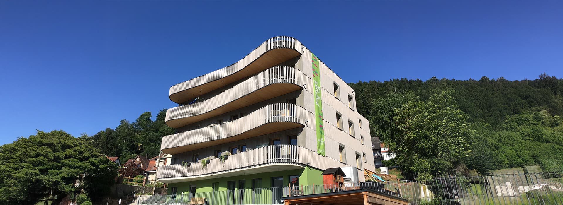 Modernes vierstöckiges Hoffnungshaus Bad Liebenzell mit geschwungenen Balkonen inmitten von Bäumen unter blauem Himmel.