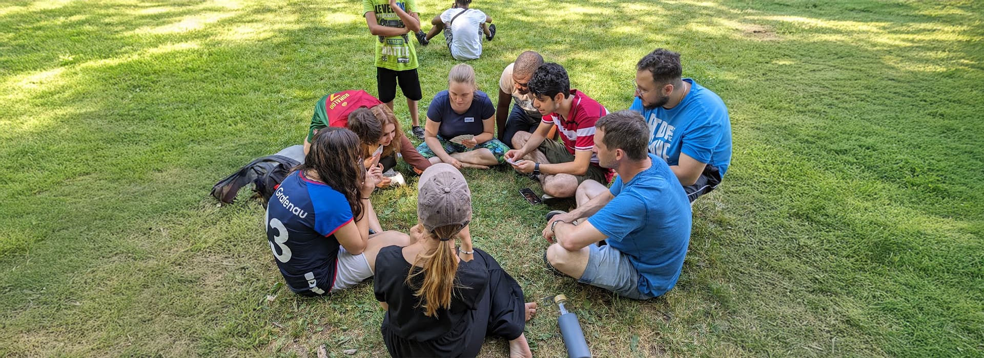 Eine Gruppe von Menschen sitzt im Kreis auf einer Wiese, diskutiert über ihre Aufgabe und konzentriert sich auf etwas in der Mitte.