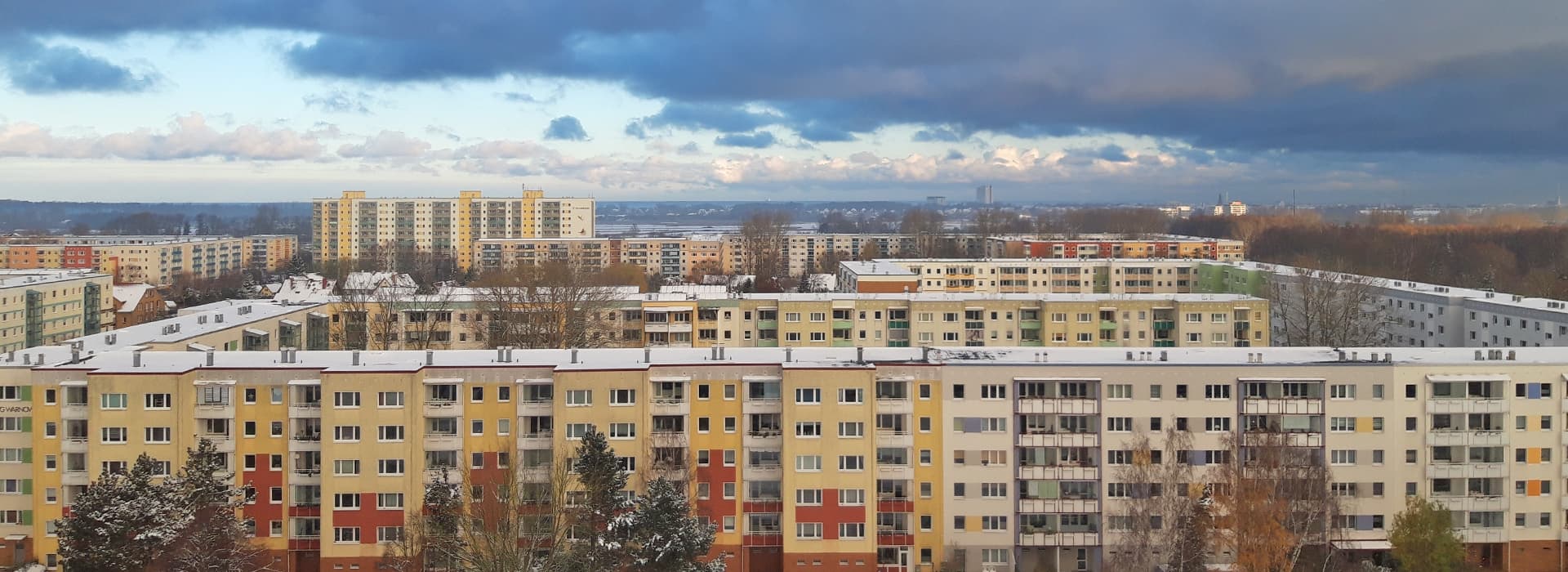 Reihen von bunten Wohnhäusern in Rostock, Deutschland, mit leichtem Schnee auf den Dächern und verstreuten Bäumen unter einem bewölkten Himmel.