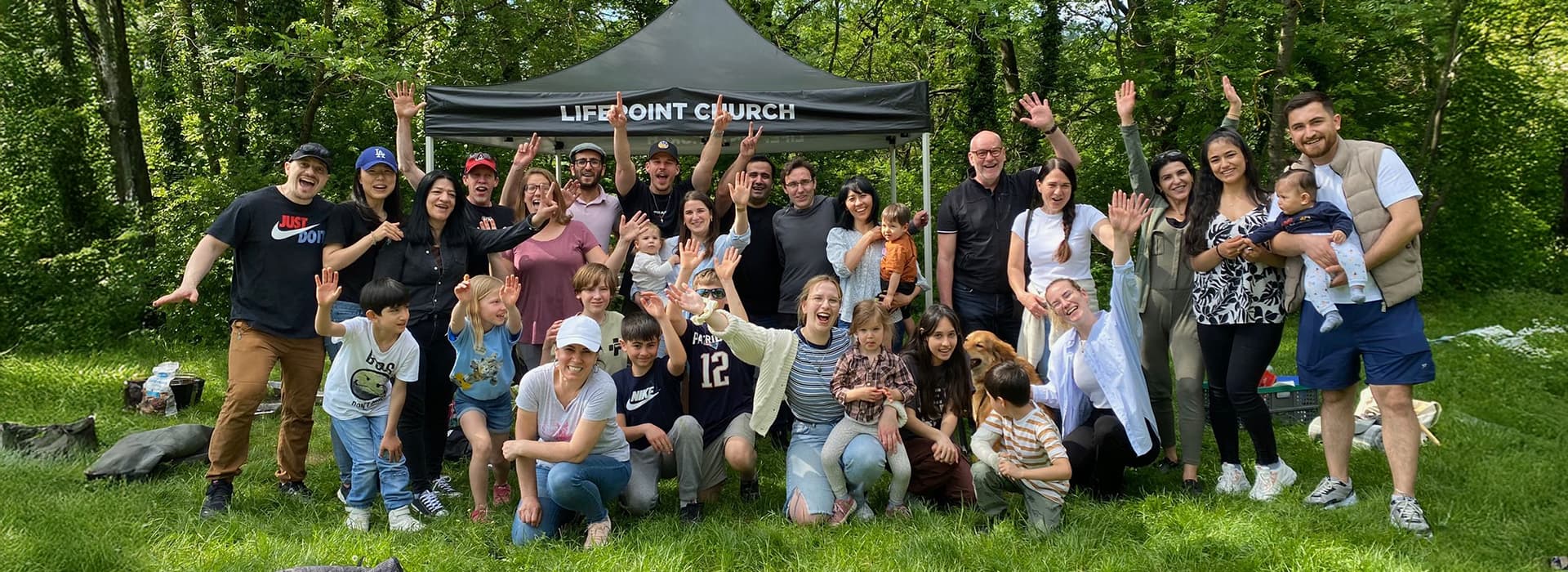 Eine bunt gemischte Gruppe lächelt und winkt vor einem Zelt der Lifepoint Church in einem Stuttgarter Park, Deutschland.