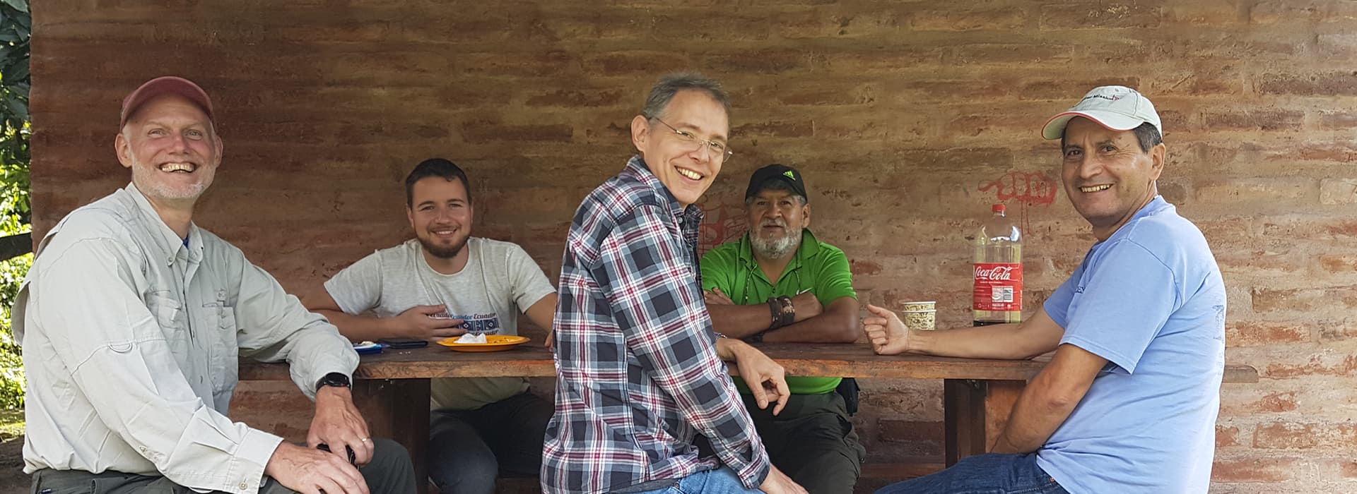 Fünf Männer sitzen nach Mitarbeiterschulungen in Ecuador um einen Holztisch im Freien, lächelnd, mit Essen und Getränken.