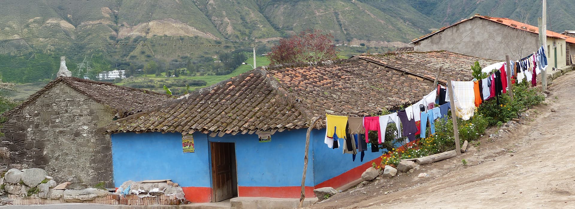 Ein blaues Haus Casa Ágape Ecuador mit Ziegeldach und bunten Kleidern auf einer Leine in einer ländlichen, bergigen Gegend.