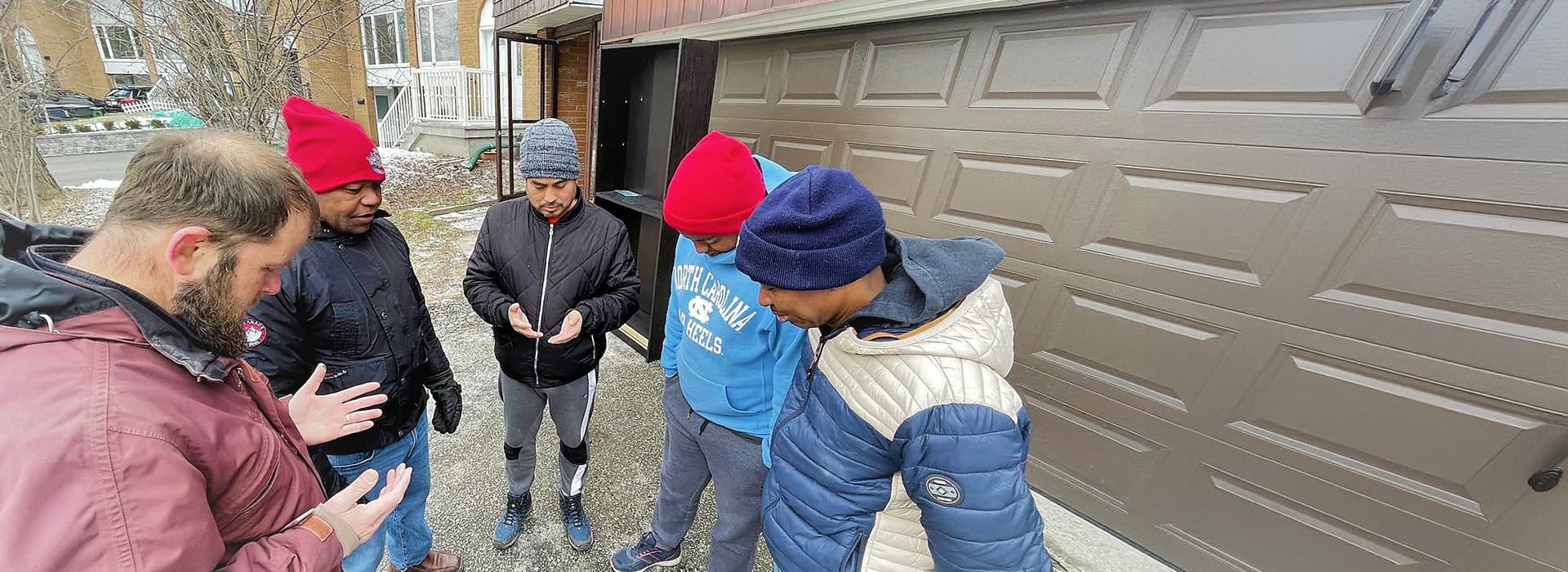 Fünf Menschen in Winterjacken und Mützen stehen betend vor einer Garage und weisen auf die Migrantenarbeit in Toronto hin.