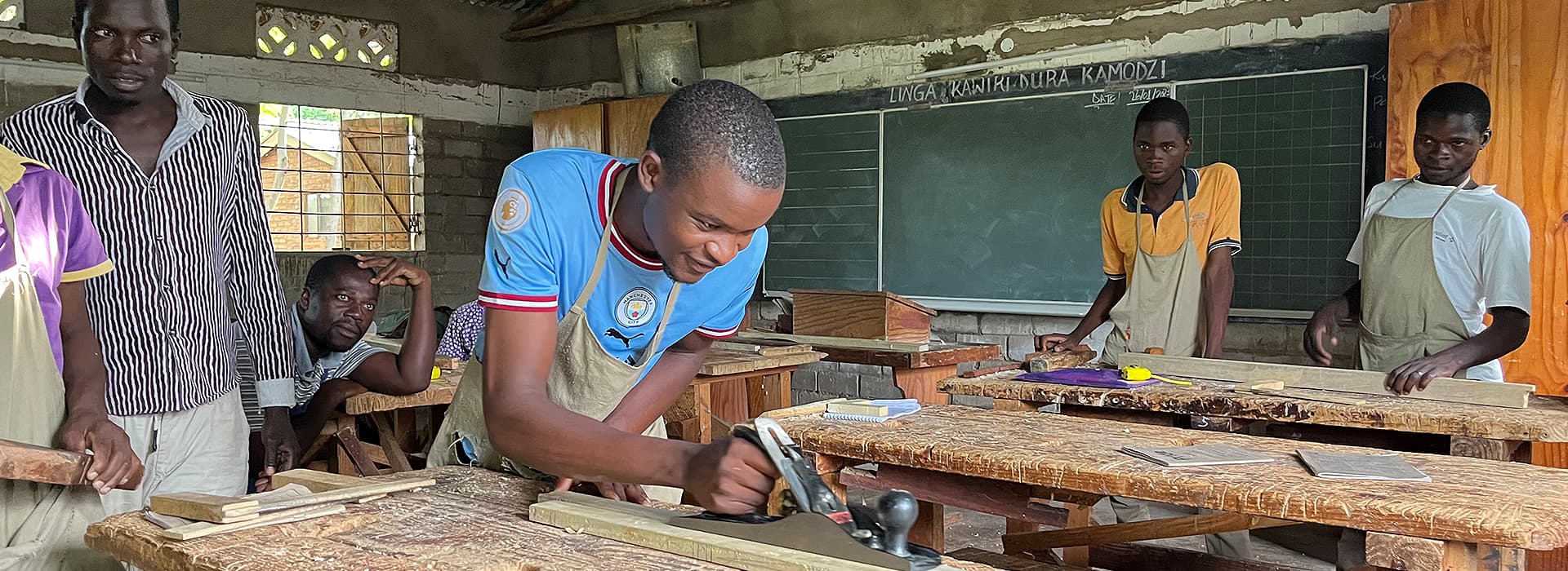 Ein Auszubildender des Chisomo-Ausbildungszentrums hobelt mit einer Handhobelmaschine Holz in einer Lehrwerkstatt, während andere zusehen.