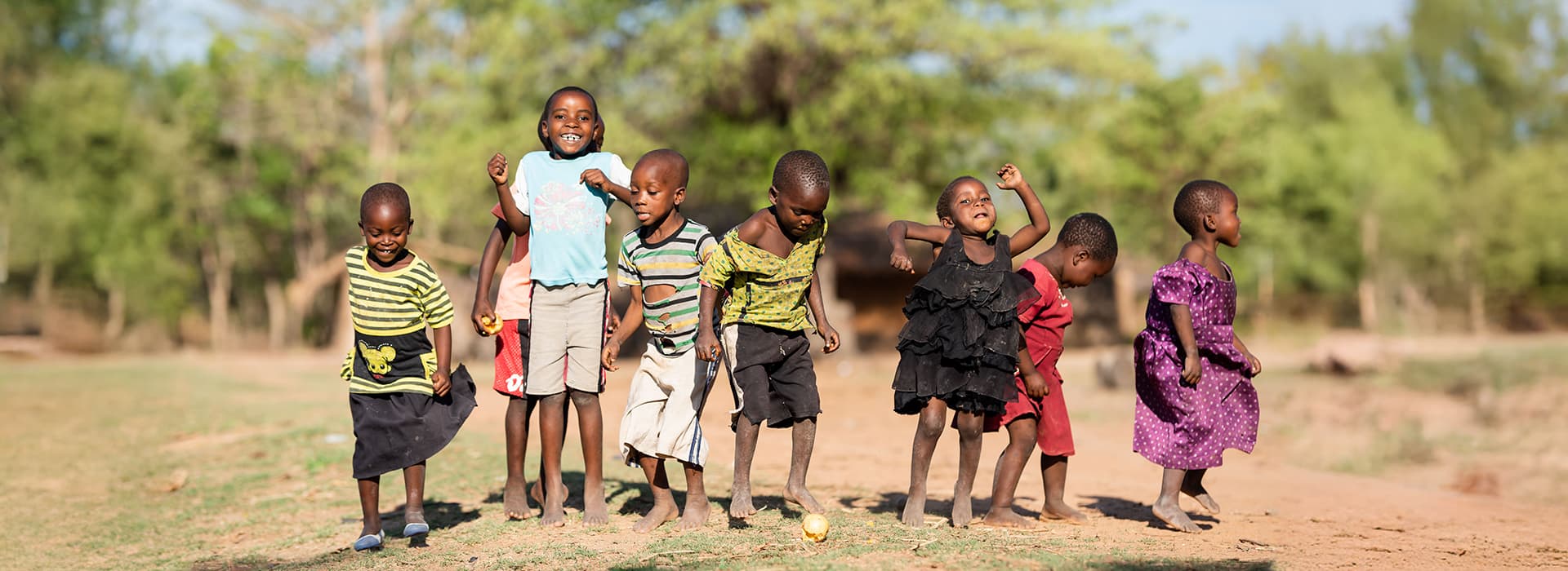 Sieben kleine Kinder in Malawi spielen während eines Dorfentwicklungsprojekts fröhlich im Freien, umgeben von Gras und Bäumen.