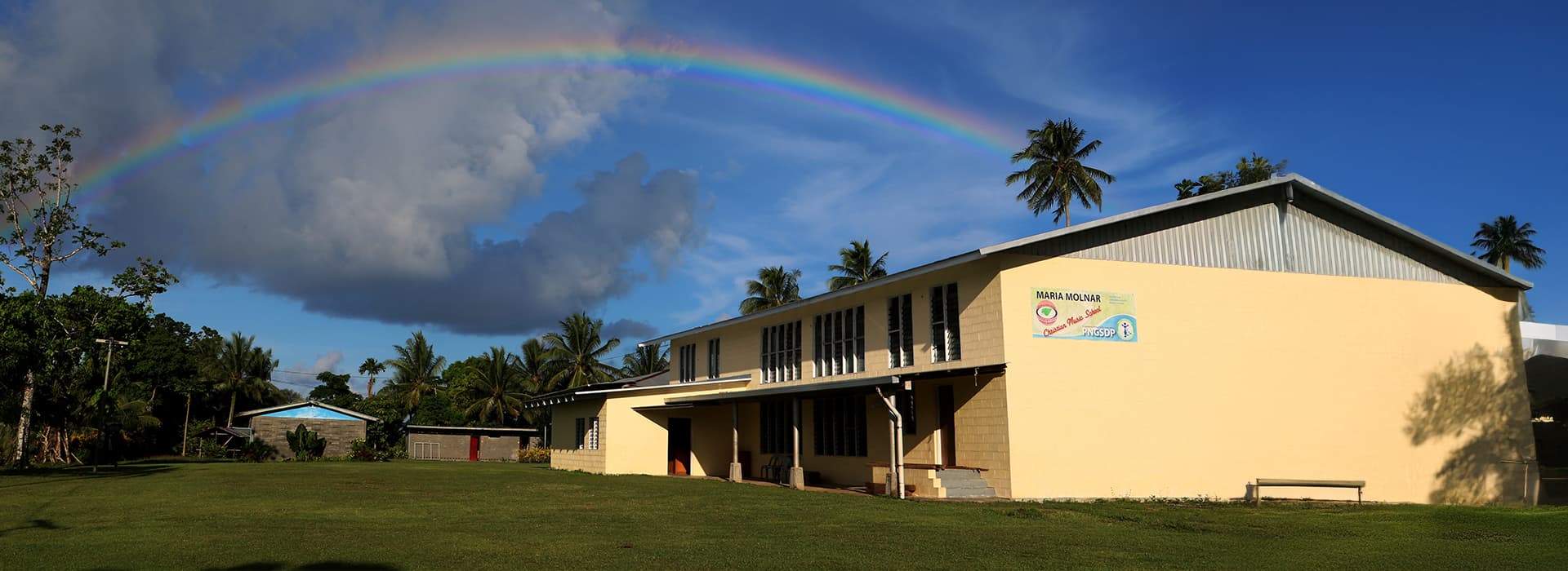 Ein gelbes Bibelschulungszentrum steht auf einer Wiese, über der sich ein Regenbogen über den teilweise bewölkten Himmel wölbt.