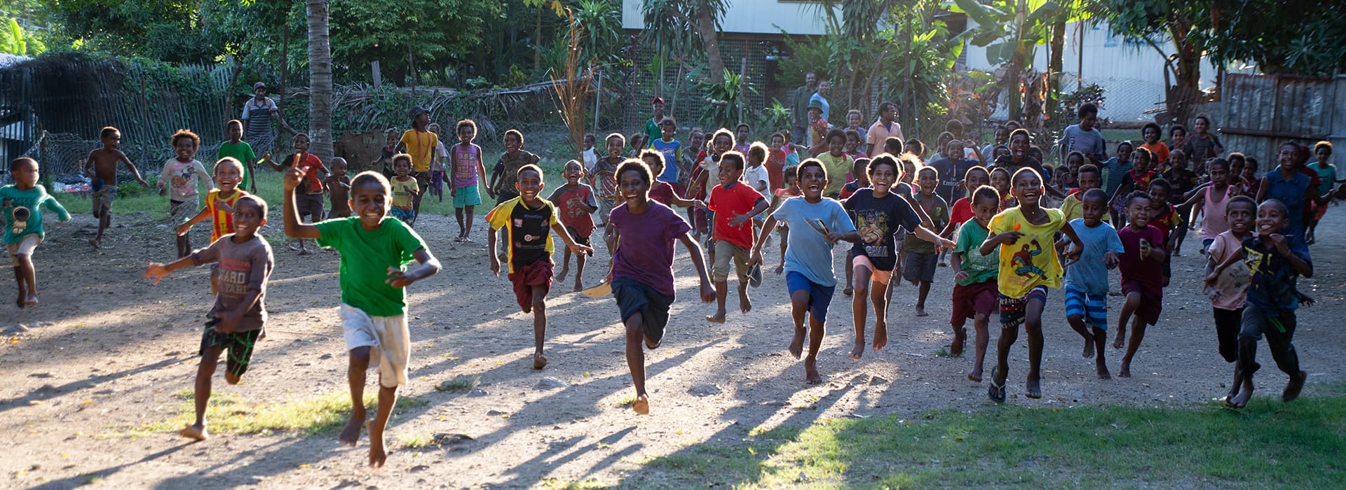 Eine große Gruppe von Kindern läuft im Freien in Papua-Neuguinea lächelnd und lachend im Sonnenlicht auf die Kamera zu.