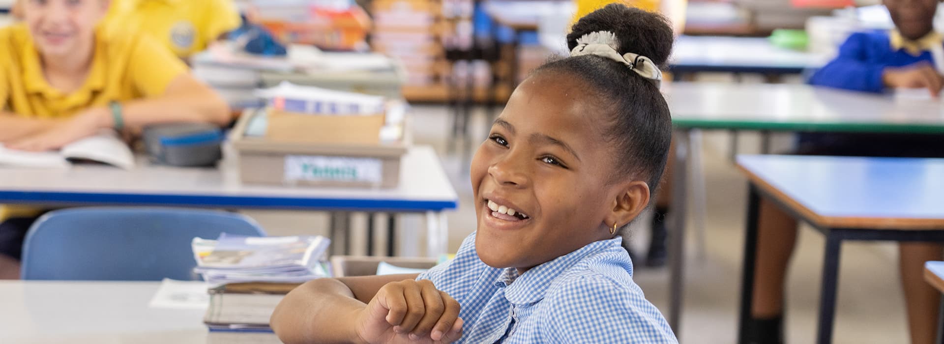 Ein junges Mädchen in einer Uniform der Amano-Schule sitzt lächelnd an ihrem Schreibtisch in einem hellen Klassenzimmer mit anderen Schülern aus Sambia.
