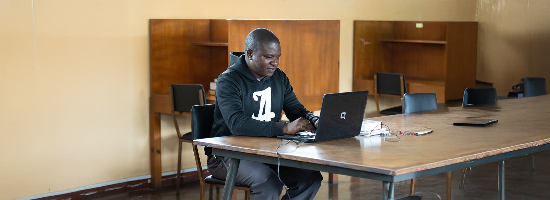 Ein Mann in einem schwarzen Kapuzenpulli arbeitet in einem ruhigen, fast leeren Raum der Evangelischen Universität in Sambia an einem Laptop.