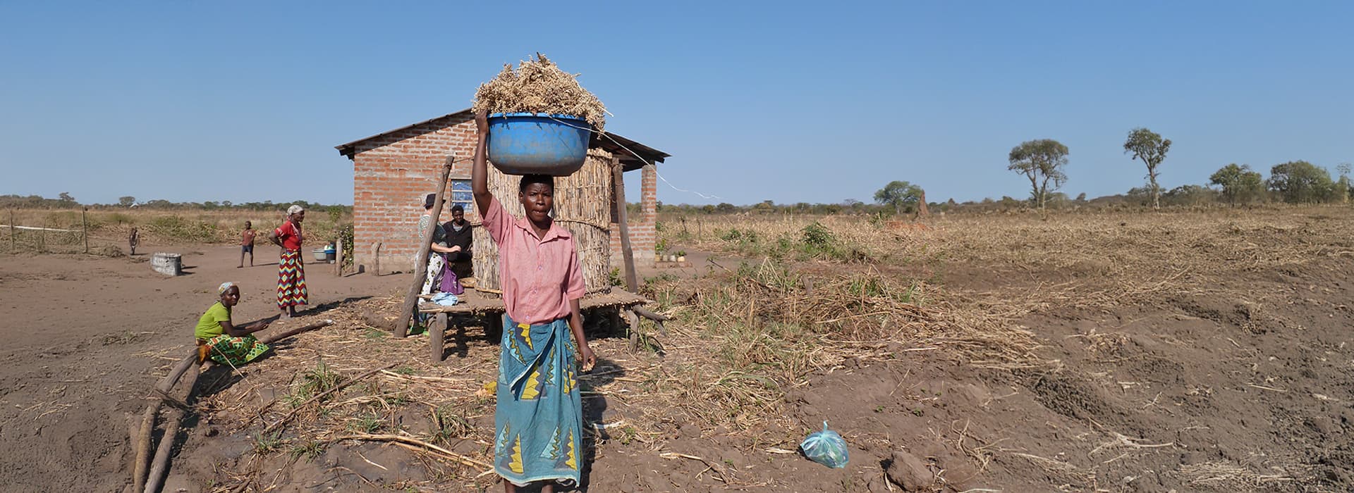 Eine Frau in Nabwalya trägt eine blaue Schüssel auf dem Kopf in der Nähe eines Backsteinhauses, in dessen Nähe sich Felder und andere Hilfesuchende befinden.