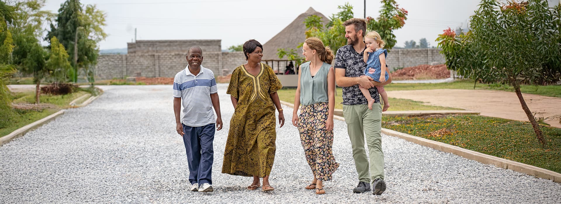Fünf Erwachsene und ein Kind in Sambia gehen lächelnd und redend auf einem Kiesweg in der Nähe von Gebäuden spazieren - Teil der Aktivitäten von Gemeindegründungen.