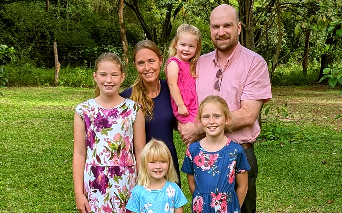 Die sechsköpfige Familie Aaron Anderson posiert in farbenfroher Kleidung auf einer Wiese mit Bäumen und Grünpflanzen im Hintergrund.