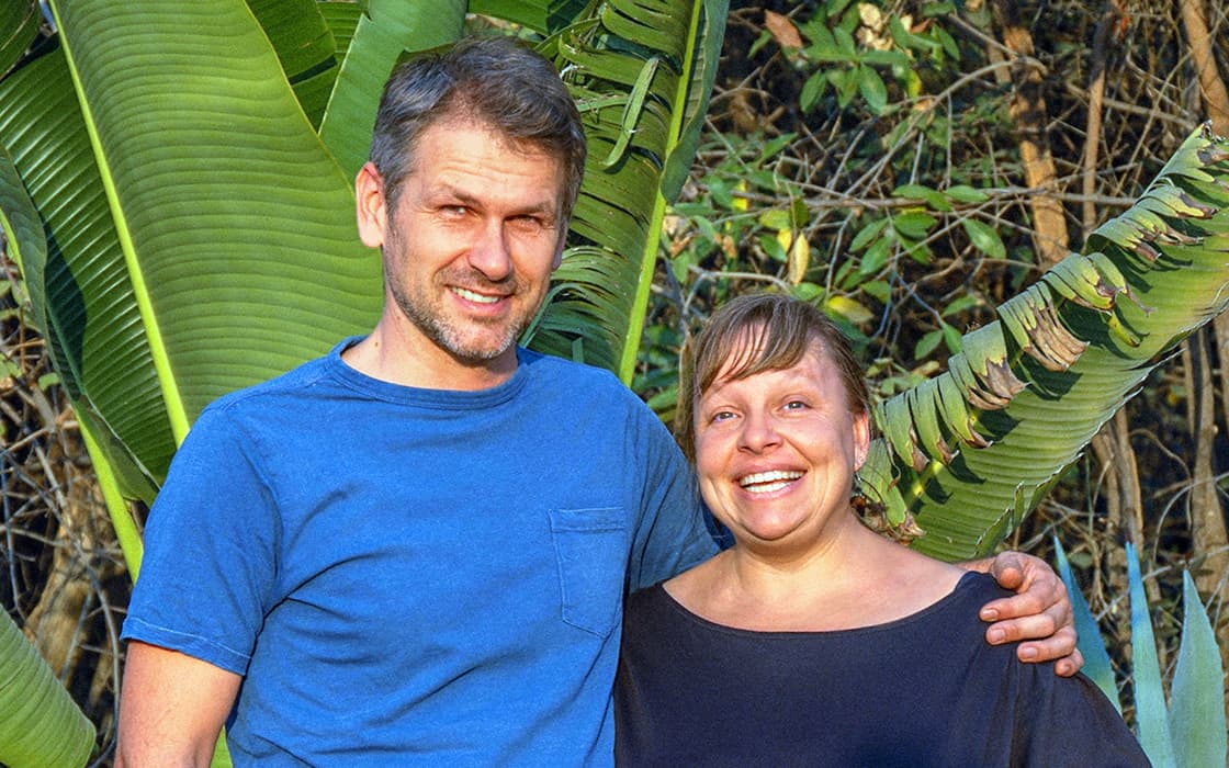 Lächelnd stehen Katja Bachmann und Matthias Bachmann gemeinsam vor großen grünen tropischen Blättern im Freien.