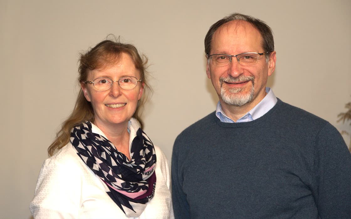 Irene Beck und Thomas Beck, lächelnde Erwachsene, stehen Seite an Seite vor einem schlichten Hintergrund.