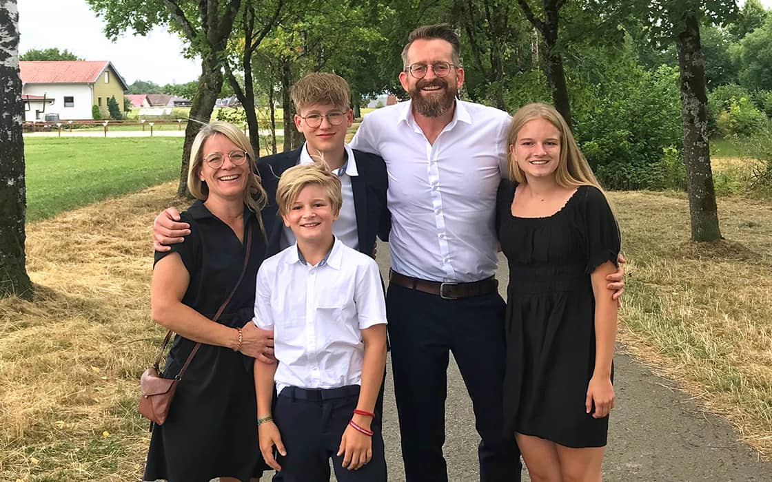 Eine fünfköpfige Familie steht lächelnd auf einem von Bäumen gesäumten Weg im Freien, mit Kerstin Böker und Joachim Böker in halb-formeller Kleidung.