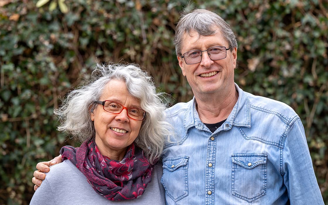 Ein lächelndes älteres Ehepaar, Katrin und Dietmar Brunner, steht mit einer Brille vor der Tür, im Hintergrund Grünpflanzen.