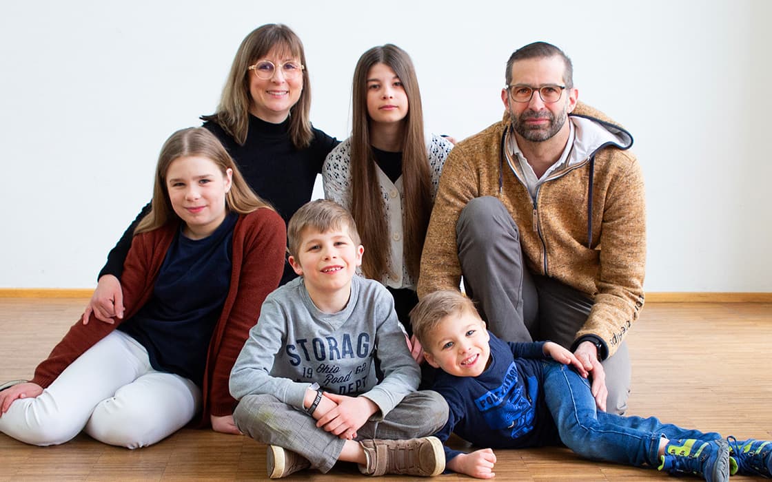 Eine sechsköpfige Familie, darunter Steffen Cramer und Melanie Cramer, lächelt auf einem Holzboden vor einem weißen Hintergrund.