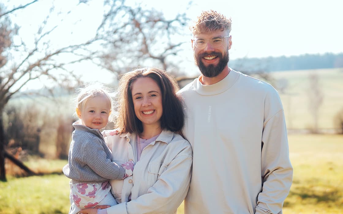 Eine lächelnde dreiköpfige Familie steht auf einer sonnigen Wiese im Freien, Niklas Ebert und Judith Ebert zwischen Bäumen und Hügeln.