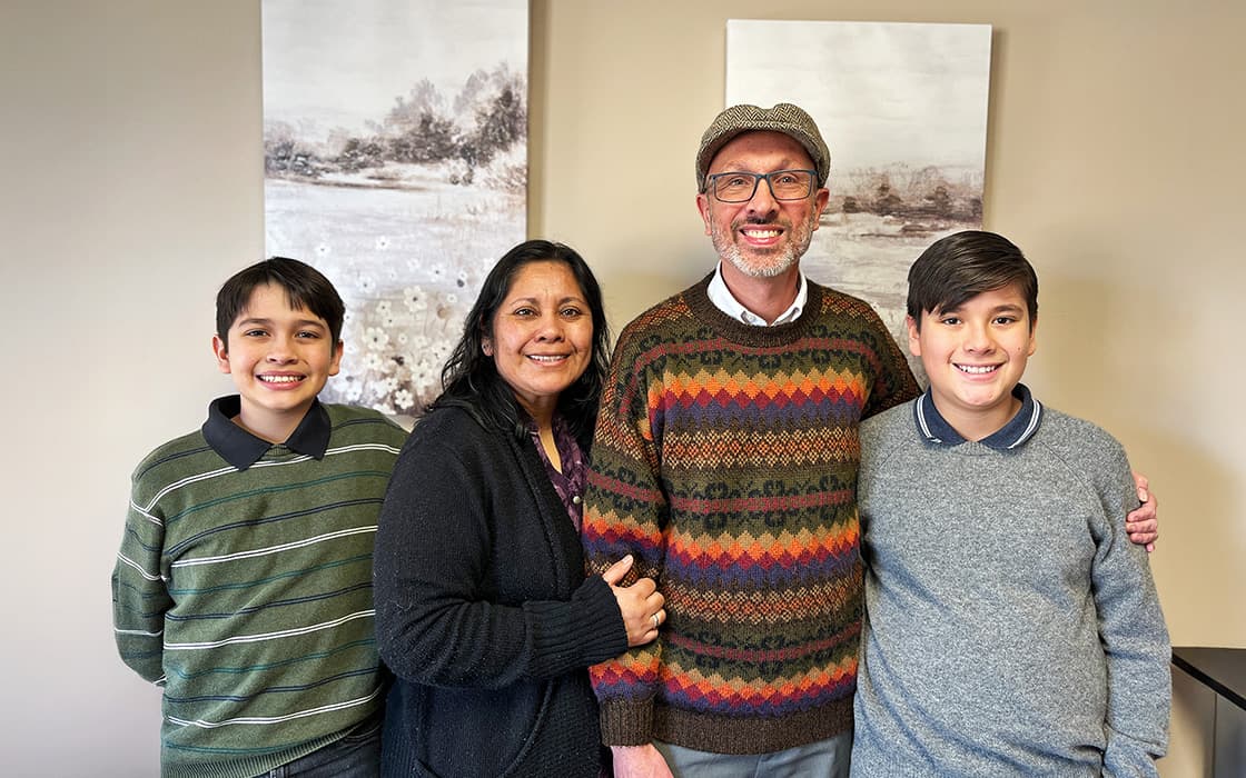 Vier Personen, darunter Ned und Marisol Farnsworth, stehen lächelnd in einem Raum vor Landschaftsgemälden mit zwei Kindern.