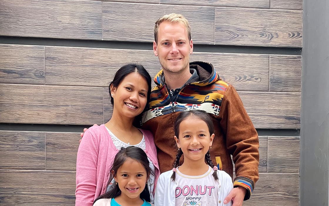 Eine lächelnde vierköpfige Familie, darunter Faith Frieß und Marcel Frieß, posiert gemeinsam vor einer Holzwand.