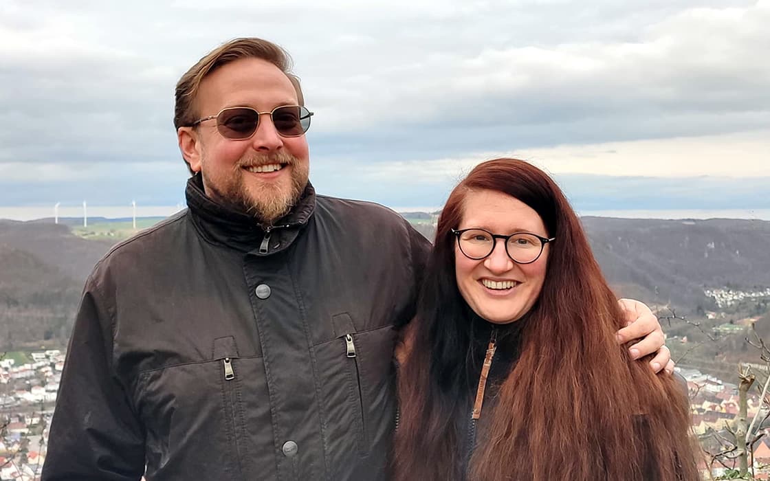 Ruth Hermann und Manuel Hermann, beide Brillenträger, lächeln im Freien mit Hügeln und einem bewölkten Himmel im Hintergrund.