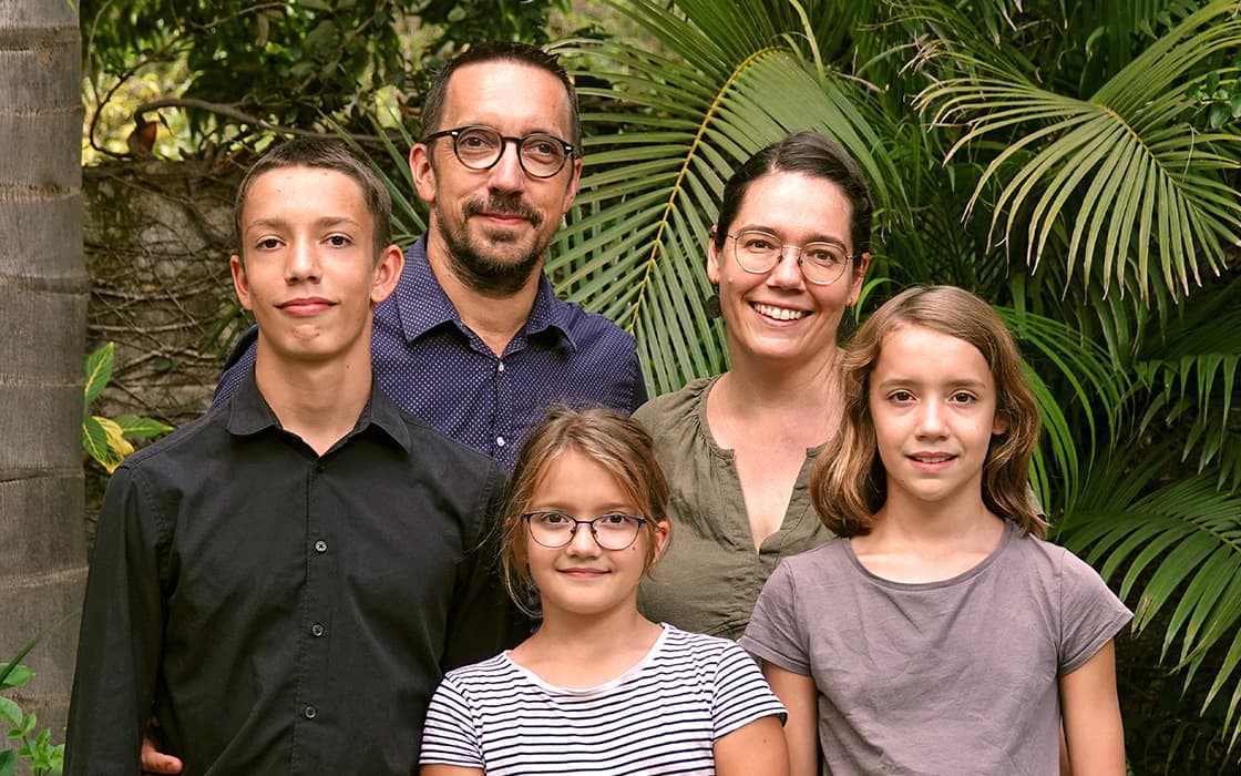 Hans-Peter Hertler und Britta Hertler posieren mit ihrer Familie im Freien, lächelnd inmitten üppiger grüner Pflanzen und Bäume.