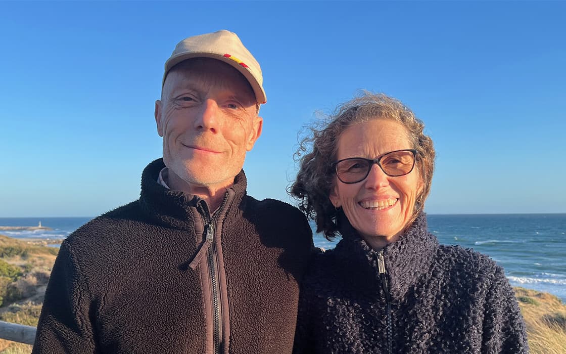Zwei lächelnde Erwachsene, Theo Hertler und Carolin Hertler, in Jacken stehen am Meer mit blauem Himmel und Strand im Rücken.