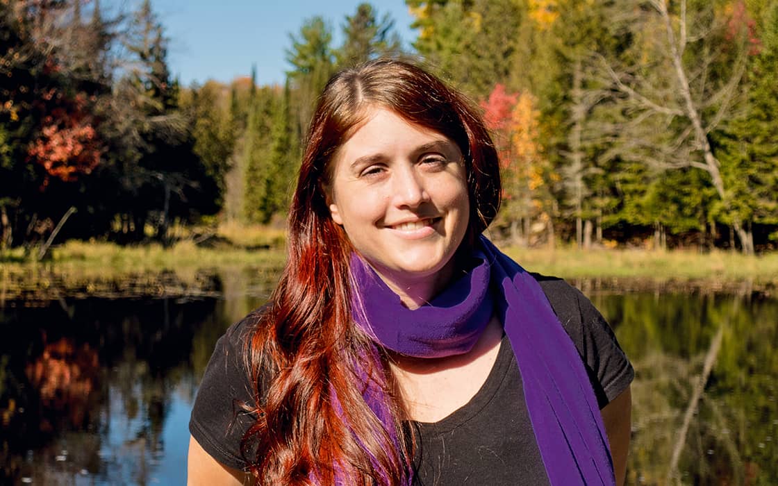 Katiana König, eine lächelnde Frau mit langen roten Haaren und einem lila Schal, steht an einem Teich mit Bäumen im Hintergrund.