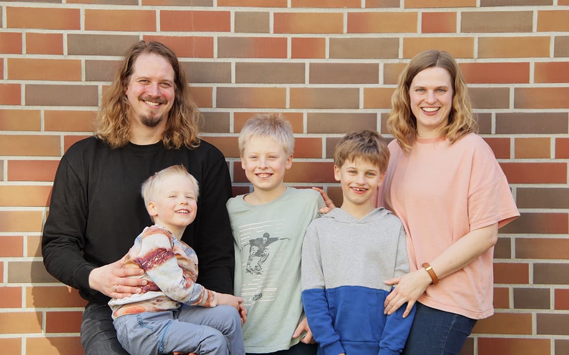 Eine lächelnde fünfköpfige Familie, darunter Aaron Köpke und Ilonka Köpke, posiert mit ihren drei Söhnen vor einer Backsteinmauer.