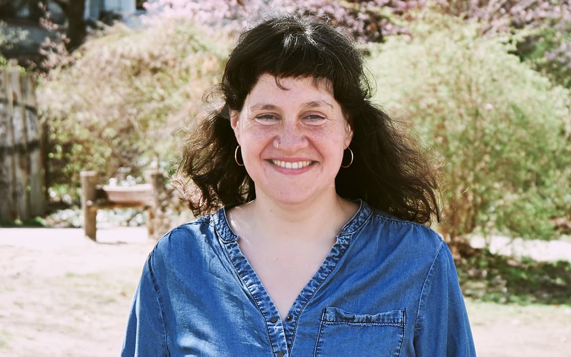 Jana Kontermann, mit dunklen Haaren, lächelt im Freien in einem blauen Jeanshemd, Bäume und eine Bank im Hintergrund.