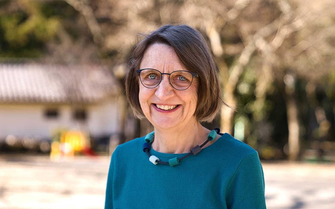 Schwester Priscilla Kunz, eine lächelnde Frau mit Brille und kurzen braunen Haaren, steht im Freien und trägt einen türkisfarbenen Pullover.