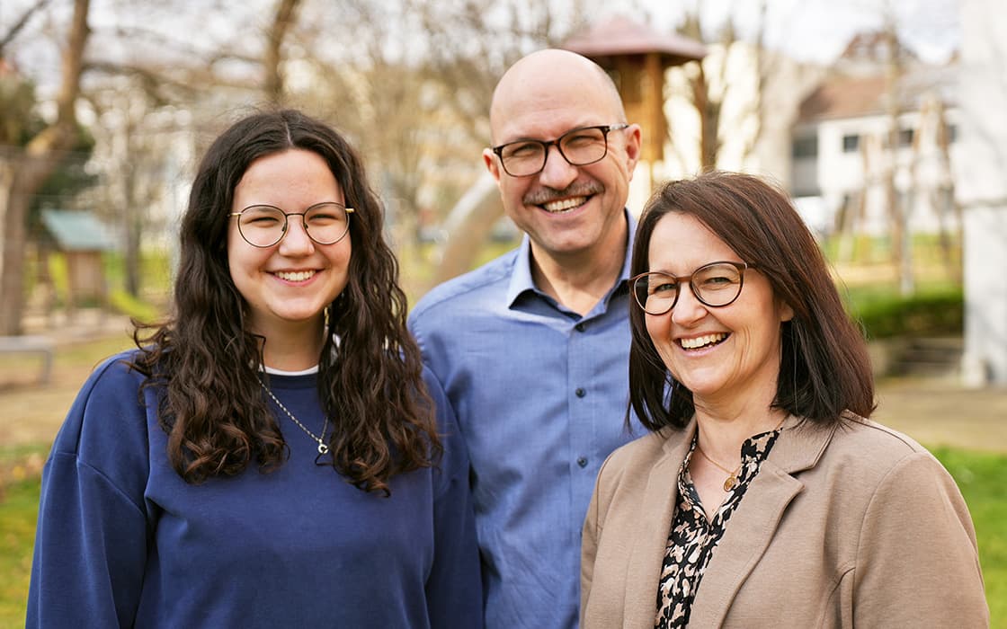 Olga Lies, Waldemar und ein Freund mit Brille lächeln gemeinsam in einer parkähnlichen Umgebung mit Bäumen im Hintergrund.
