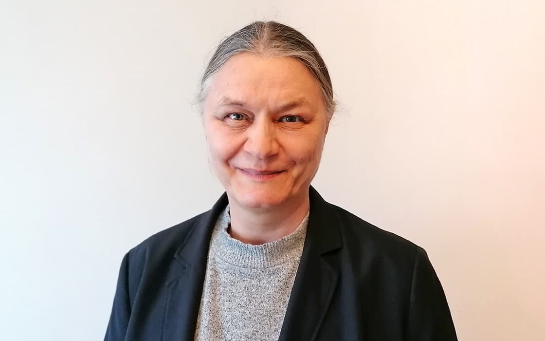 Lächelnde Schwester Sabine Matthis mit grauem Haar in einem Dutt, trägt einen grauen Pullover und einen schwarzen Blazer, vor einem hellen Hintergrund.