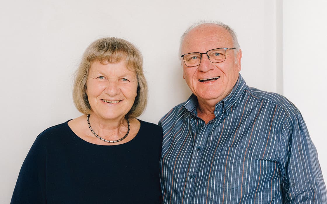 Ein lächelndes älteres Ehepaar, Klaus-Dieter Mauer und Ulrike Mauer, steht mit Brille und Freizeitkleidung vor einer weißen Wand.
