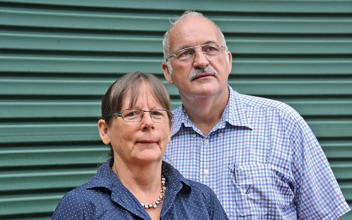 Irmgard Mortsiefer und Bernd Mortsiefer, beide mit Brille und Kragenhemd, stehen vor einer grünen Wellblechwand.