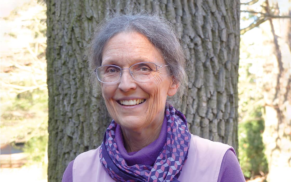 Eine lächelnde ältere Frau, Dr. Esther Pflaum, mit grauem Haar und Brille, steht draußen vor einem Baum und trägt einen lila Schal.