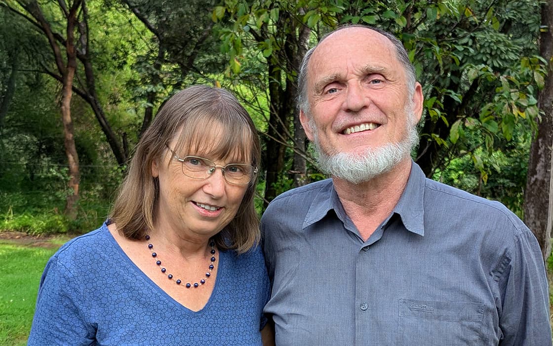 Bärbel Pflaum und Michael Pflaum stehen lächelnd zusammen im Freien mit Bäumen und Grünzeug im Hintergrund.